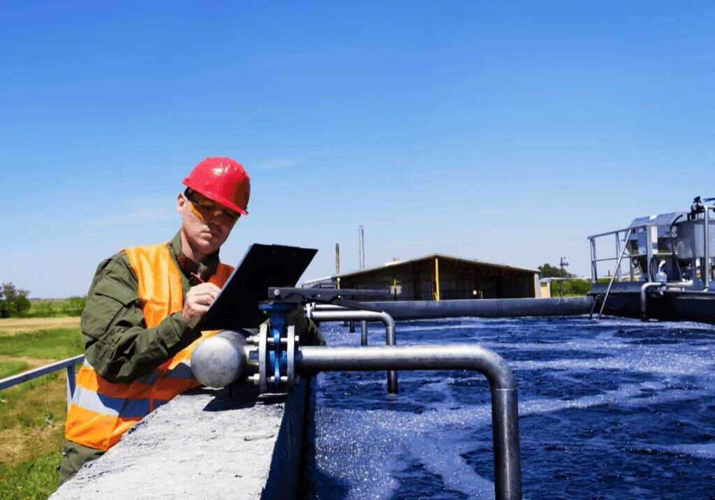 A man in an orange hat and yellow vest sitting on the side of a river.